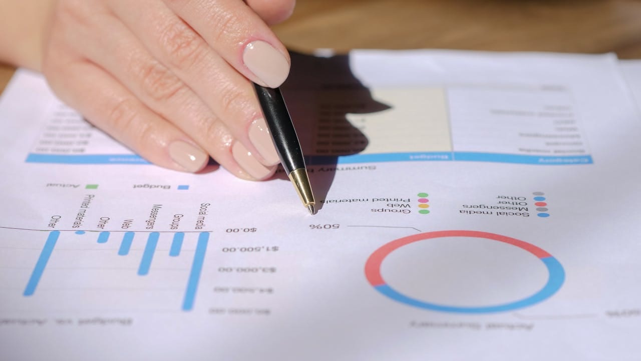 service-05 Close-up of a hand with a pen pointing at a financial document with graphs and charts.